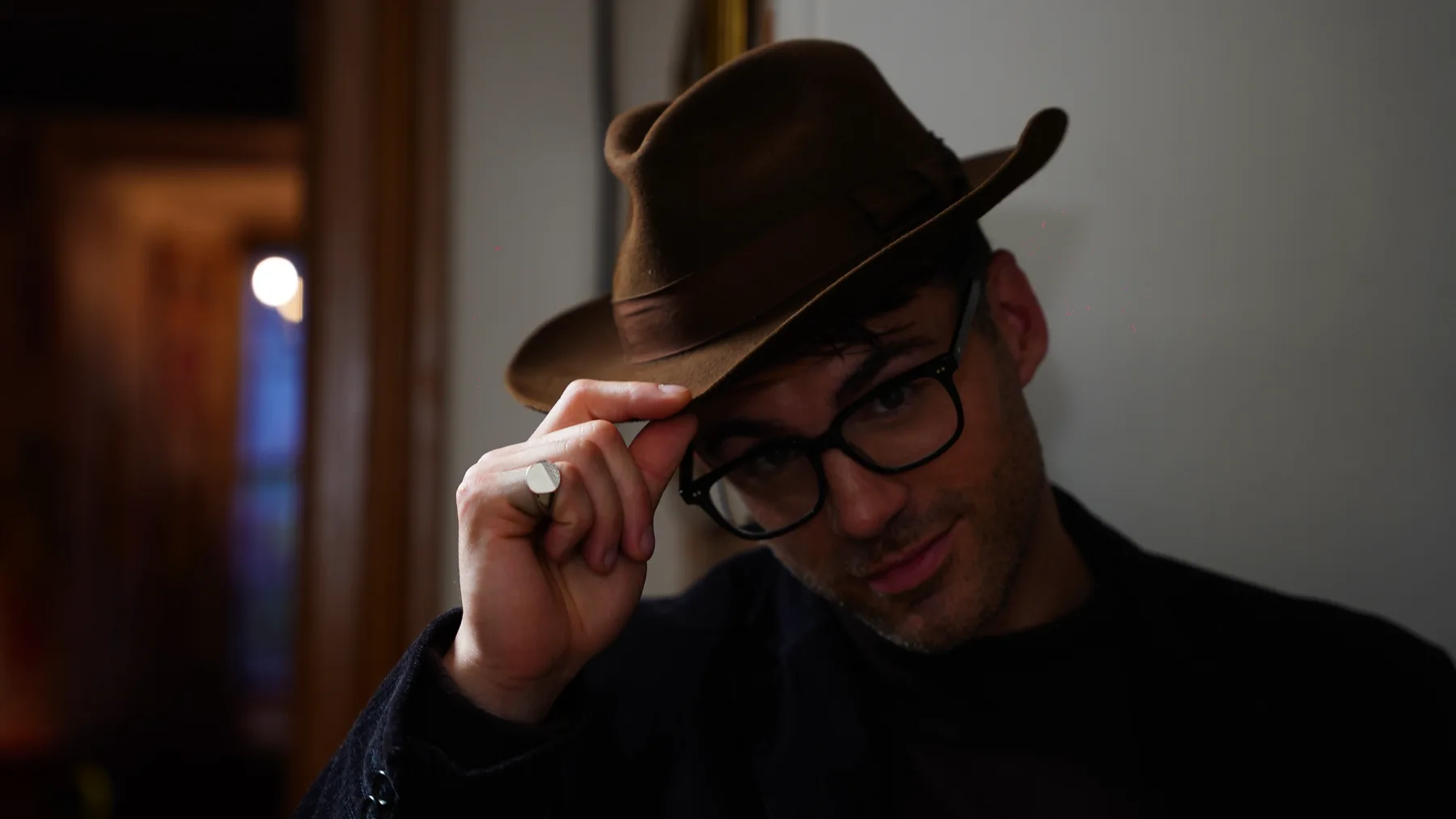 A man in a brown hat and glasses tips his hat in a dim interior on the Murder at Macintosh Manor set
