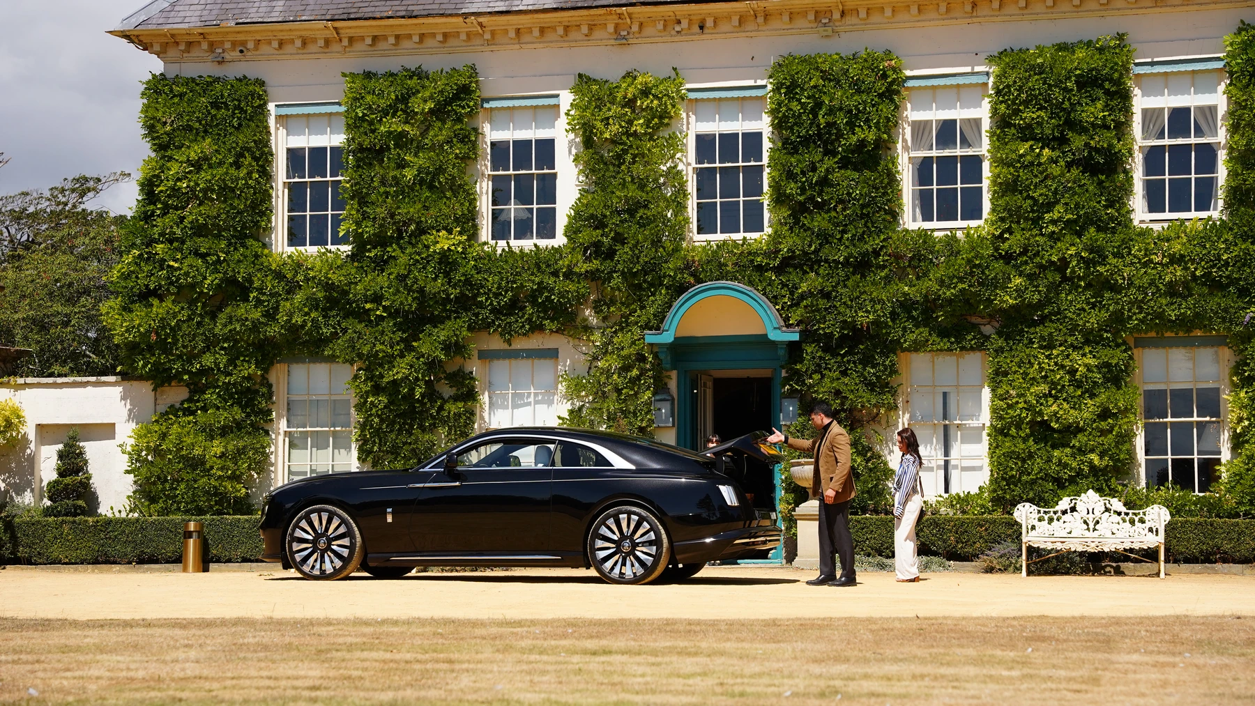 A black car and two cast members outside the ivy-covered manor house