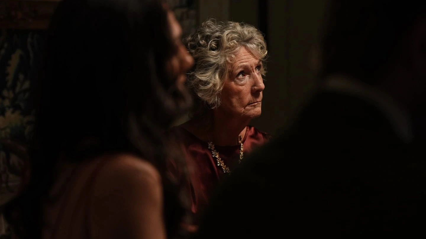 Dark, low-lit still of an older woman with curled grey hair and a pearl necklace, gazing upward while blurred figures crowd the foreground