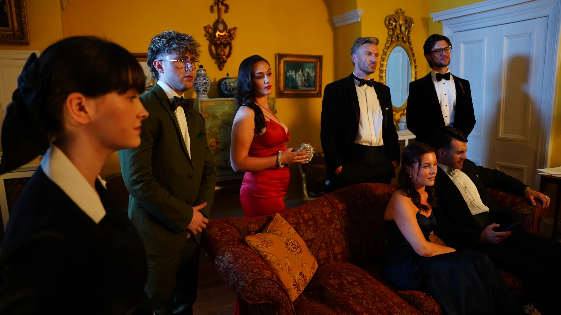 Still of a group in evening wear gathered in a mustard-yellow drawing room, some standing and two seated on a red patterned sofa