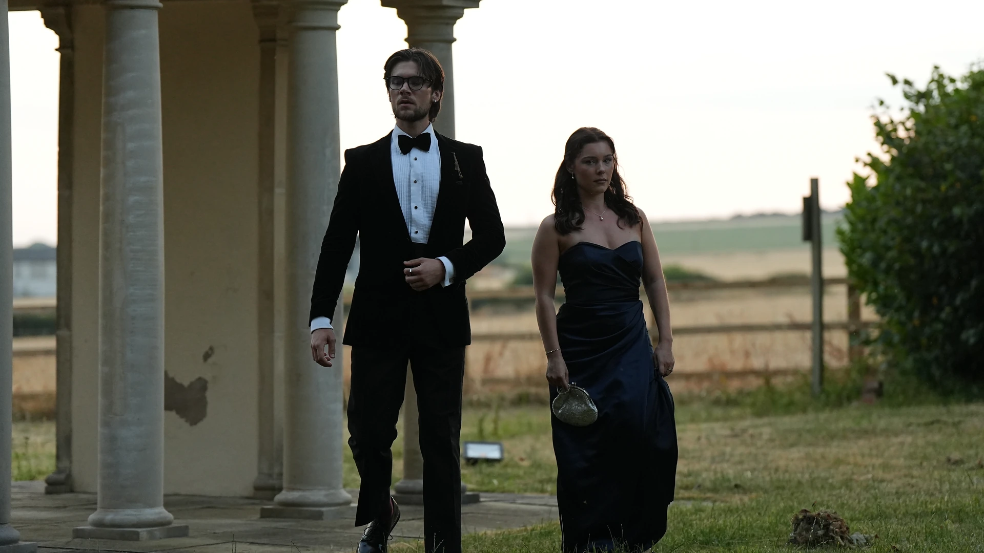 Still of a man in a dinner suit and bow tie and a woman in a navy gown walking past stone columns onto a lawn at dusk